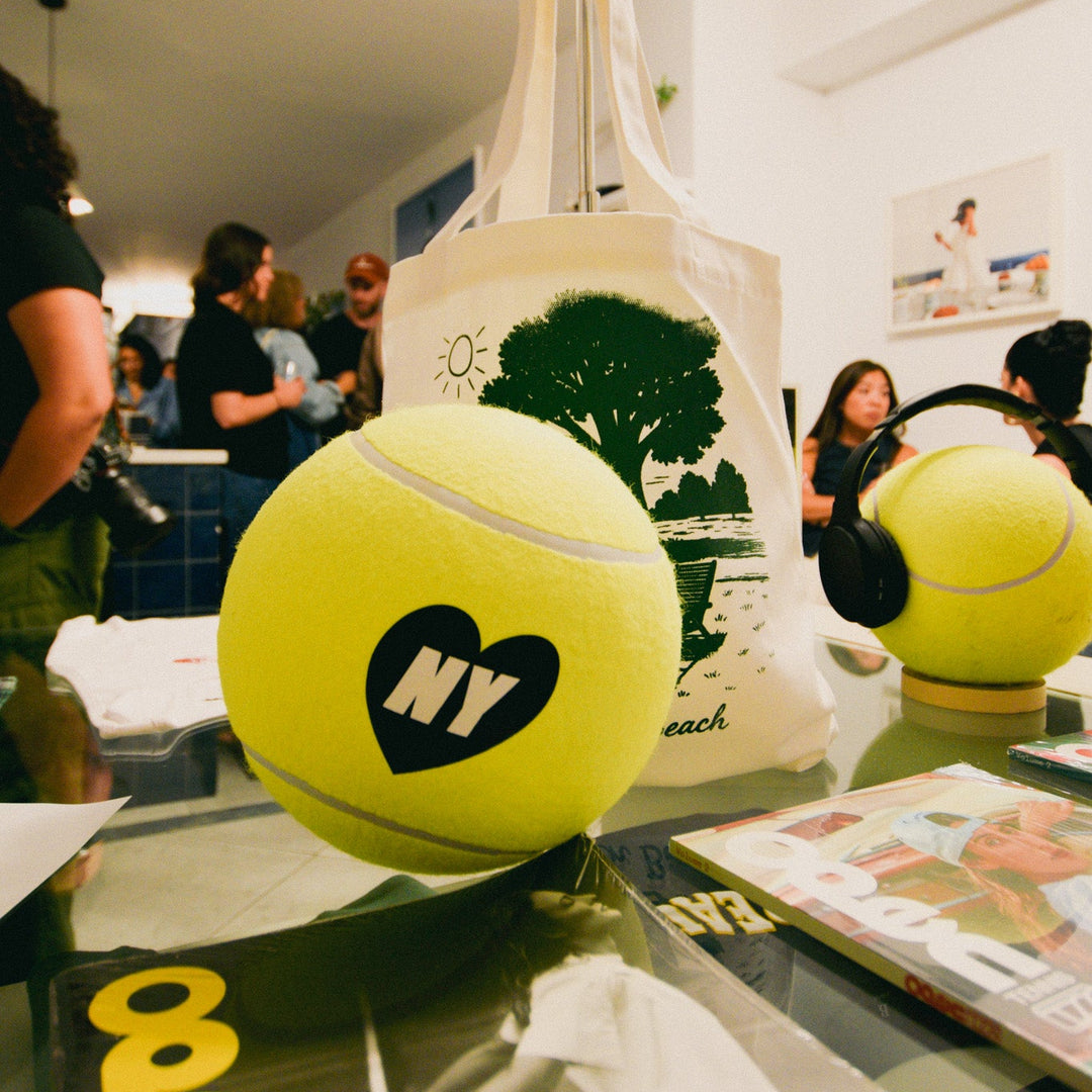Two yellow tennis balls with 'NY' and a heart logo on a glass table, with people in the background.