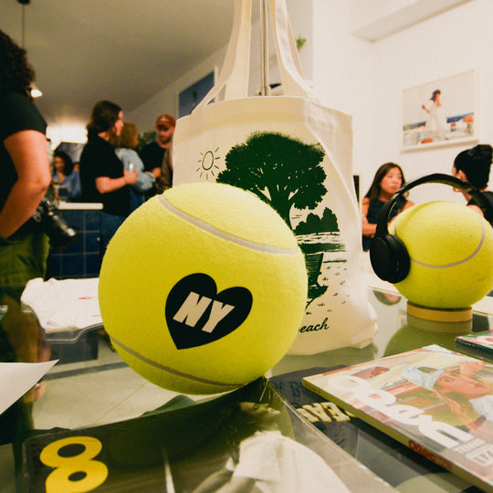 Two yellow tennis balls with 'NY' and a heart logo on a glass table, with people in the background.
