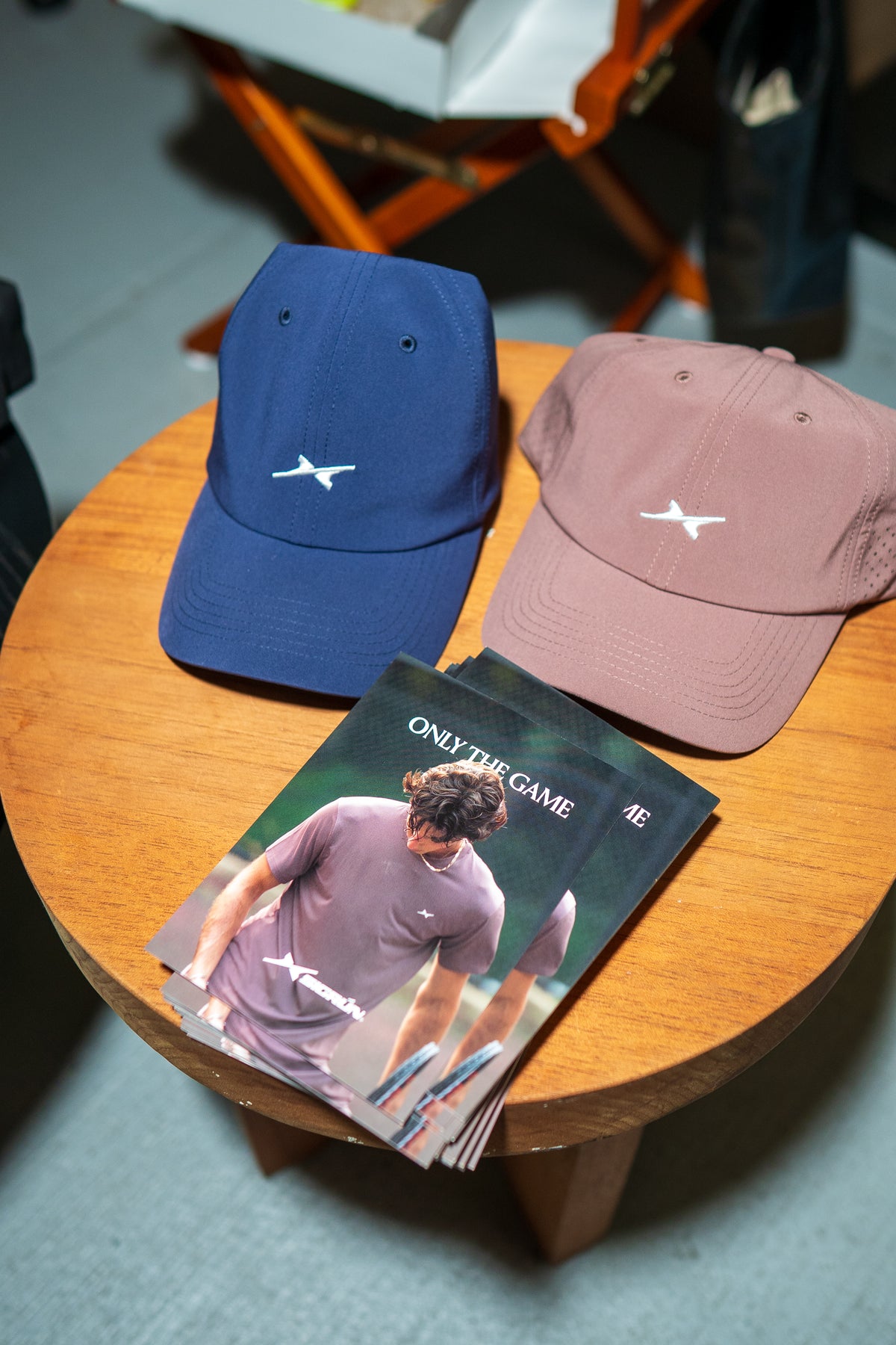 Two baseball caps, one navy blue and one dusty rose pink, displayed on a round wooden table next to a folded magazine. Both caps feature a small white embroidered logo on the front.